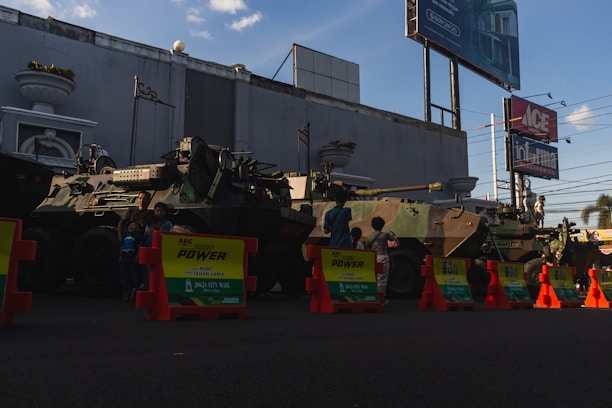 Two military armored vehicles are parked in a row on a street, partially obscured by orange and yellow traffic barriers in front. Several children stand around and climb on the vehicles. The background features a large building with minimalistic decoration, billboards, and power lines. The sky is clear with a few clouds.