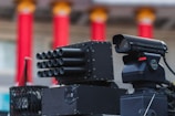A close-up view of advanced, military-style equipment that appears to be a surveillance or targeting system. The setup includes multiple cylindrical components suggesting a multi-barrel design and an adjacent black device resembling a camera or sensor with a lens. The background features red, vertical columns with orange tops, indicating an architectural setting.