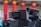 A close-up view of advanced, military-style equipment that appears to be a surveillance or targeting system. The setup includes multiple cylindrical components suggesting a multi-barrel design and an adjacent black device resembling a camera or sensor with a lens. The background features red, vertical columns with orange tops, indicating an architectural setting.
