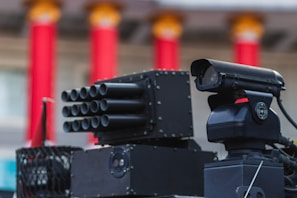 A close-up view of advanced, military-style equipment that appears to be a surveillance or targeting system. The setup includes multiple cylindrical components suggesting a multi-barrel design and an adjacent black device resembling a camera or sensor with a lens. The background features red, vertical columns with orange tops, indicating an architectural setting.