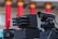 A close-up view of advanced, military-style equipment that appears to be a surveillance or targeting system. The setup includes multiple cylindrical components suggesting a multi-barrel design and an adjacent black device resembling a camera or sensor with a lens. The background features red, vertical columns with orange tops, indicating an architectural setting.