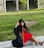 A woman wearing a graduation cap and gown is seated on a curb in a grassy park area. Her black gown features yellow initials, and she is wearing a bright red dress and black high heels. There are trees and a building in the background.