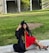 A woman wearing a graduation cap and gown is seated on a curb in a grassy park area. Her black gown features yellow initials, and she is wearing a bright red dress and black high heels. There are trees and a building in the background.