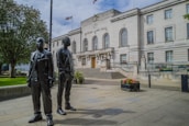 Two dark statues stand in front of a large, neoclassical-style building labeled 'Hackney Town Hall.' The building features multiple arched windows and a symmetrical facade with steps leading to the entrance. Flags are flying atop the building. The foreground includes paved walkways, potted plants with colorful flowers, and a well-maintained lawn with trees.
