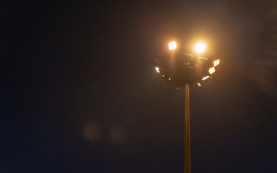 A tall streetlight is illuminated against a dark night sky, casting a warm glow. The light fixtures create a halo-like effect in the night, emphasizing the contrast between light and darkness.