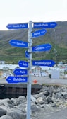 A directional signpost with multiple blue signs pointing towards various global cities and locations including South Pole, North Pole, London, Beijing, Canberra, Berlin, Paris, and Ólafsfjörður. The background features a mountainous landscape, a communications tower, a small harbor with rocks, and buildings in a quaint town.