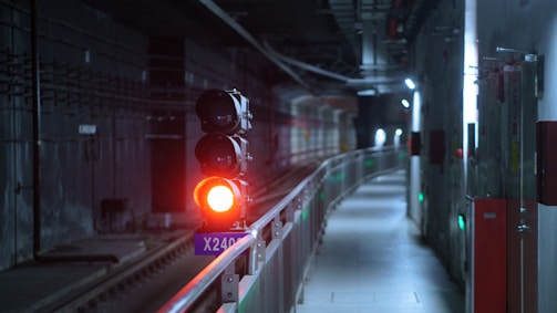 An underground tunnel with a train signal displaying a red light. The tunnel is dimly lit, with metallic railings and panels on the walls. The environment appears industrial with a focus on infrastructure.
