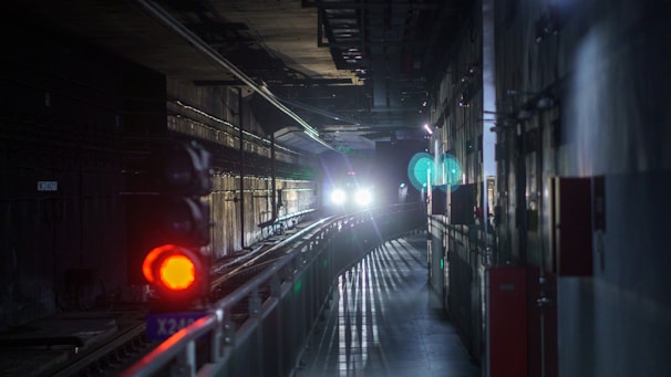 A dimly lit underground tunnel with a train approaching. Bright headlights illuminate the tracks ahead, casting shadows on the walls. A red light signal is visible on the left, indicating a stop or caution signal. The walls are lined with various cables and equipment.