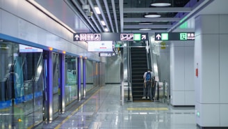 A spacious and modern subway station platform with clean, shiny floors. Illuminated directional signs hang overhead, guiding passengers to escalators leading upwards. A person stands on the escalator, moving to the upper level. The platform is lined with clear glass barriers next to the train tracks.