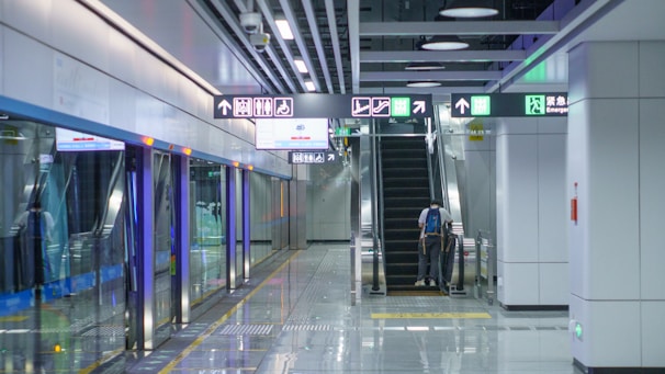 A spacious and modern subway station platform with clean, shiny floors. Illuminated directional signs hang overhead, guiding passengers to escalators leading upwards. A person stands on the escalator, moving to the upper level. The platform is lined with clear glass barriers next to the train tracks.