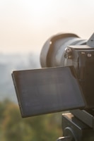 A digital camera with a large lens is positioned to take a photo, with its screen displaying camera settings. Soft sunlight is illuminating the scene against a blurred natural backdrop.