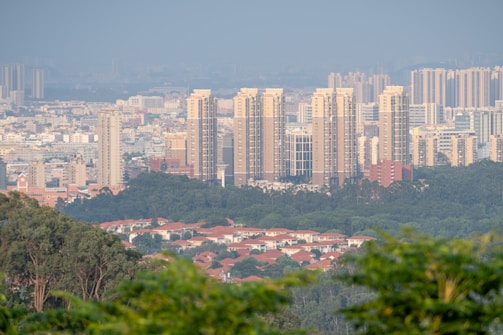 A cityscape featuring tall, modern apartment buildings with a backdrop of shorter, densely packed structures. In the foreground, a lush green area with scattered trees frames rows of red-roofed houses.