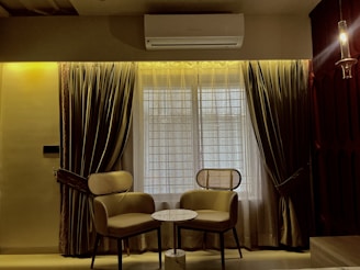 Two chairs with wicker backs are positioned around a small round marble table in front of a large window framed by heavy curtains. An air conditioner is mounted above the window, and a hanging light fixture is visible on the right side. The room has a warm ambiance with muted lighting.