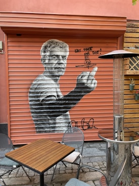 A large mural of a man is painted on a metal shutter. The artwork is monochrome, contrasting with the orange background. The figure is gesturing with one hand extended forward. Graffiti text next to the mural reads 'Eat, Taste or Die!'. In the foreground, there is a small outdoor seating area with a wooden table, metal chairs, and a tall outdoor heater.