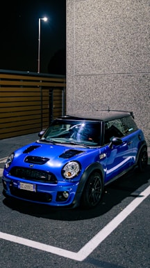 A blue Mini Cooper is parked in an outdoor setting at night, illuminated by a nearby streetlight. The car is positioned in a parking space near a tall, textured building wall. The asphalt pavement and parking lines are clearly visible, adding to the urban setting.