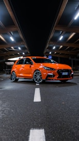 A bright orange hatchback car is parked in a dimly lit, covered parking area. The car has a sporty and modern design, featuring sleek lines and alloy wheels. Fluorescent lights above create reflections on the car's surface, enhancing its vibrant color.