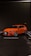 An orange sports car is parked in an outdoor urban setting, with wet pavement reflecting its vibrant color. The car features sleek, aerodynamic design and alloy wheels, positioned in a marked parking spot against a dark, tiled wall.