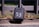 A black lock box with the number 53 written on it in white is secured to a metal gate. The box is positioned against a stone surface, with a grassy area visible in the blurred background.