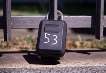 A black lock box with the number 53 written on it in white is secured to a metal gate. The box is positioned against a stone surface, with a grassy area visible in the blurred background.