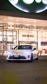 A sleek, white sports car is parked in front of a modern building at night. The building has large glass doors and is well-lit, with the words 'POGLIANO MILANESE' and 'INGRESSO' displayed above. The car's headlights are on.