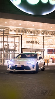 A sleek, white sports car is parked in front of a modern building at night. The building has large glass doors and is well-lit, with the words 'POGLIANO MILANESE' and 'INGRESSO' displayed above. The car's headlights are on.
