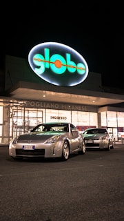 Two silver sports cars are parked in front of a brightly lit store entrance at night. The storefront features a large, illuminated sign with the word 'globo' and the location 'Pogliano Milanese'. The scene is well-lit, highlighting the sleek design of the cars.