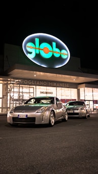 Two silver sports cars are parked in front of a brightly lit store entrance at night. The storefront features a large, illuminated sign with the word 'globo' and the location 'Pogliano Milanese'. The scene is well-lit, highlighting the sleek design of the cars.