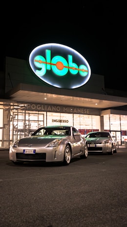Two silver sports cars are parked in front of a brightly lit store entrance at night. The storefront features a large, illuminated sign with the word 'globo' and the location 'Pogliano Milanese'. The scene is well-lit, highlighting the sleek design of the cars.