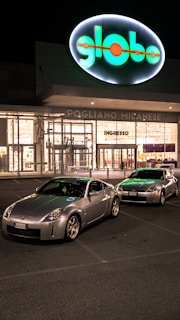Two silver sports cars are parked in front of a brightly lit shopping center at night. The building has a large, illuminated sign with the word 'globo' and an entrance labeled 'ingresso'. The parking lot is relatively empty, and the scene conveys a quiet, serene atmosphere.