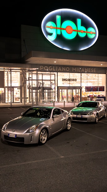 Two silver sports cars are parked in front of a brightly lit shopping center at night. The building has a large, illuminated sign with the word 'globo' and an entrance labeled 'ingresso'. The parking lot is relatively empty, and the scene conveys a quiet, serene atmosphere.