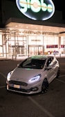 A sleek white car with prominent front lights is parked in front of a brightly lit modern building at nighttime. The building has large glass windows and a sign reading 'Pogliano Milanese.' The glowing brand sign, 'globo,' is prominently displayed above the entrance.