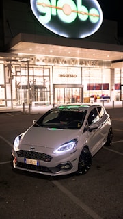 A sleek white car with prominent front lights is parked in front of a brightly lit modern building at nighttime. The building has large glass windows and a sign reading 'Pogliano Milanese.' The glowing brand sign, 'globo,' is prominently displayed above the entrance.