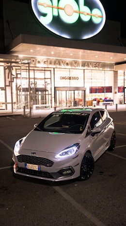 A sleek white car with prominent front lights is parked in front of a brightly lit modern building at nighttime. The building has large glass windows and a sign reading 'Pogliano Milanese.' The glowing brand sign, 'globo,' is prominently displayed above the entrance.