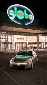 A silver sports car is parked in front of a brightly lit storefront at night. The store's sign, illuminated in green and orange, casts a reflection on the car's hood. The storefront has large glass windows, displaying goods inside. The location appears to be a commercial zone with clear parking lines on the asphalt.