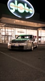 A silver sports car with a European license plate is parked in front of a shopping center at night. The building behind has a large illuminated sign that reads 'globo'. The atmosphere is calm with minimal activity around.