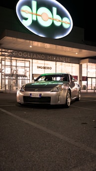 A silver sports car with a European license plate is parked in front of a shopping center at night. The building behind has a large illuminated sign that reads 'globo'. The atmosphere is calm with minimal activity around.