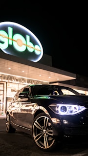 A sleek black car is parked in front of a well-lit commercial building with a neon 'globo' sign. The car's headlights are on, creating a striking reflection on its polished surface.