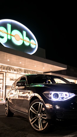 A sleek black car is parked in front of a well-lit commercial building with a neon 'globo' sign. The car's headlights are on, creating a striking reflection on its polished surface.