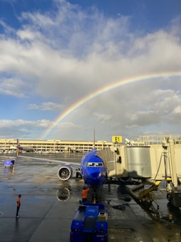 A vibrant rainbow arches across a partly cloudy sky above an airport tarmac. Below, a blue airplane is stationed at gate E1, connected to a jet bridge. Two airport workers in orange vests are visible on the wet pavement, possibly managing ground operations.