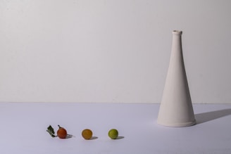 A minimalist composition features a white conical vase on the right side and three small fruits of varying colors—red, orange, and green—on the left against a plain background. A couple of green leaves lie next to the red fruit, and the overall setup is simple and balanced.