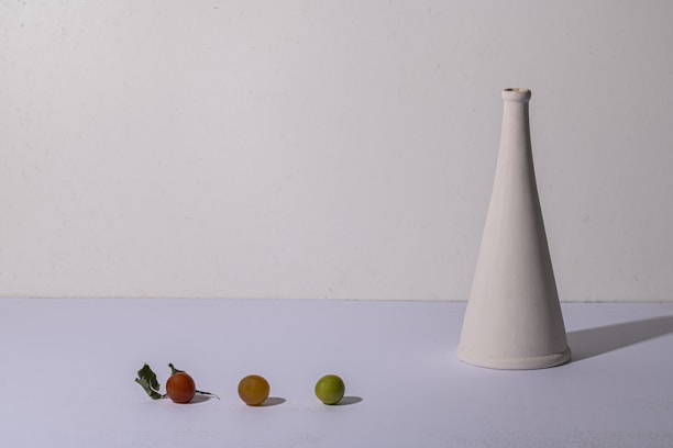 A minimalist composition features a white conical vase on the right side and three small fruits of varying colors—red, orange, and green—on the left against a plain background. A couple of green leaves lie next to the red fruit, and the overall setup is simple and balanced.