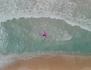 Aerial view of a person on a pink surfboard navigating through the waves of a beach shoreline. The waves create white foam against the teal ocean water, with a sandy beach below.