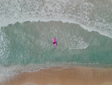 Aerial view of a person on a pink surfboard navigating through the waves of a beach shoreline. The waves create white foam against the teal ocean water, with a sandy beach below.