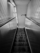 A black and white photograph of an upward-moving escalator, bordered by tiled walls. A security camera is mounted on the wall at the top, capturing a sense of surveillance.