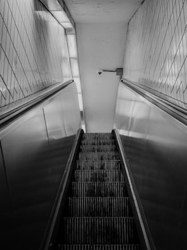 A black and white photograph of an upward-moving escalator, bordered by tiled walls. A security camera is mounted on the wall at the top, capturing a sense of surveillance.