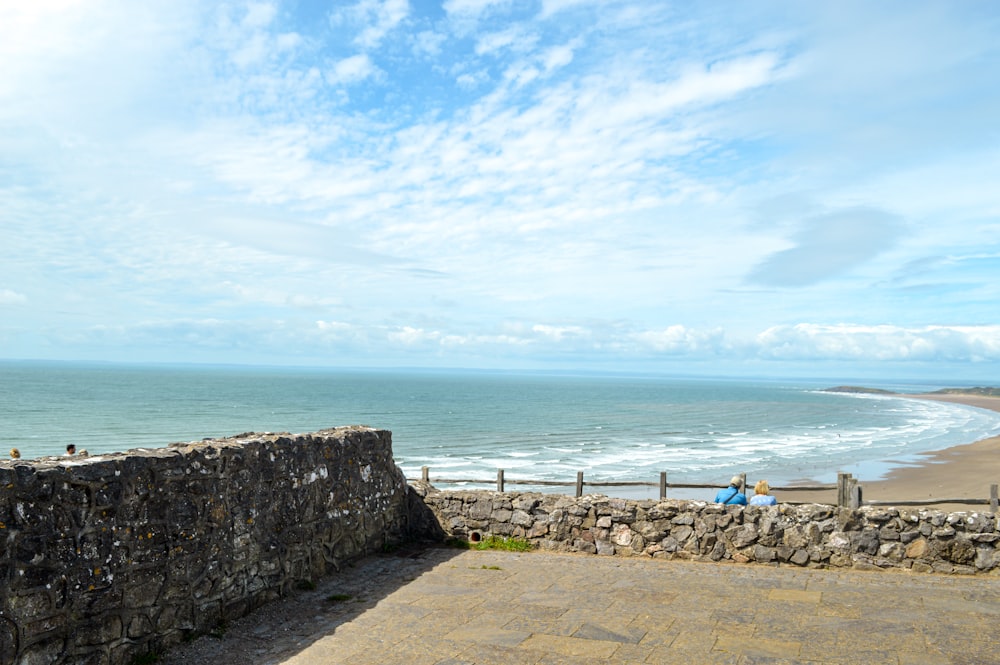 a stone wall next to the ocean with people sitting on it