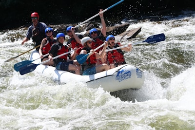 A group of people are white-water rafting down a river, wearing helmets and life jackets. They are in a white inflatable raft, navigating through turbulent waters, appearing enthusiastic and engaged in the adventure.