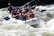 A group of people are white-water rafting down a river, wearing helmets and life jackets. They are in a white inflatable raft, navigating through turbulent waters, appearing enthusiastic and engaged in the adventure.