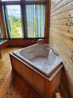 A wooden-paneled room features a white whirlpool bathtub with jets, positioned in the corner. Large windows with green curtains allow natural light to fill the room, offering views of greenery outside. The wooden floorboards complement the rustic look of the interior.
