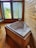 A wooden-paneled room features a white whirlpool bathtub with jets, positioned in the corner. Large windows with green curtains allow natural light to fill the room, offering views of greenery outside. The wooden floorboards complement the rustic look of the interior.
